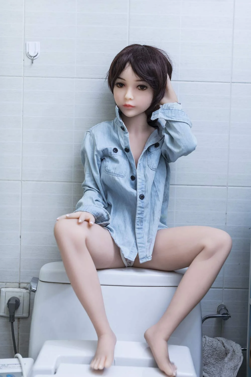 A mini sex doll sitting on the toilet seat and touching his head