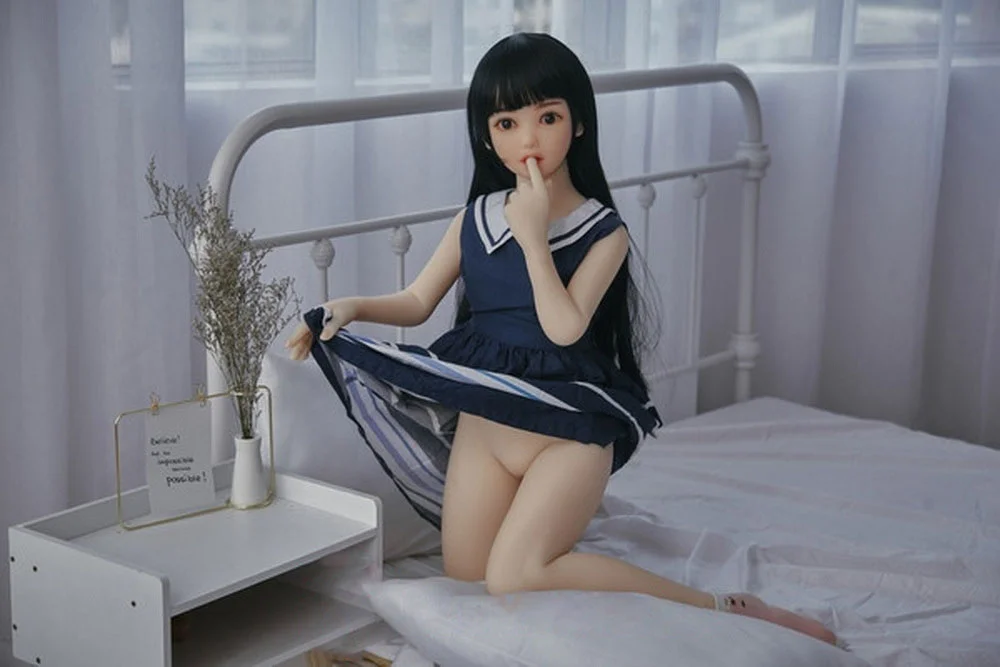 Flat-chested sex doll kneeling on the bed with fingers in mouth